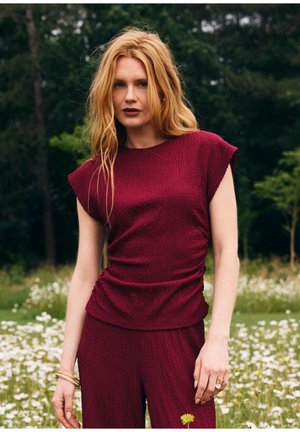 Femme aux longs cheveux blonds portant un haut sans manches texturé rouge foncé et un pantalon assorti, debout dans un champ de fleurs blanches.