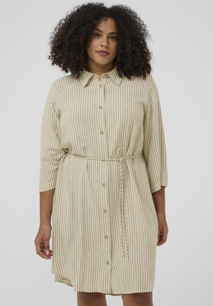 Kobieta o kręconych włosach, ubrana w beżową sukienkę w pionowe białe paski, zapinaną na guziki, z rękawami o długości 3/4 i cienkim paskiem w talii.