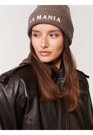 Brązowa czapka z ribbowanym splotem z napisem "LA MANIA" w białym kolorze, noszona z ciemną skórzaną kurtką. Gładka tekstura i dopasowany krój.