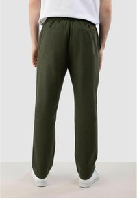 Olijfgroene, relaxed-fit broeken met een elastische tailleband, achterzakken en een gladde textuur. Gecombineerd met witte sneakers.
