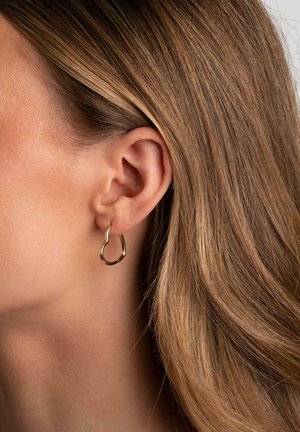 Nahaufnahme eines Ohres mit hellbraunem Haar, das einen kleinen, unregelmäßig geformten goldenen Creolenring trägt, vor einem schlichten Hintergrund.