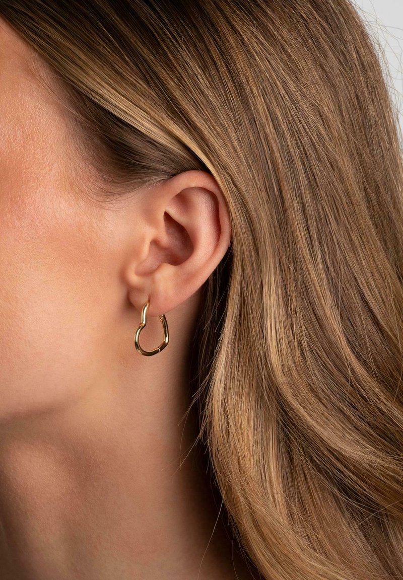 Nära bild på ett öra med ljusbrunt hår som bär en liten, oregelbundet formad guldsmycke mot en neutral bakgrund.