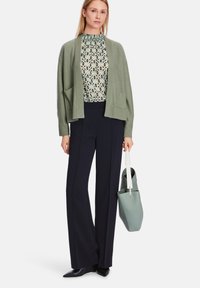 Hellgrüne Strickjacke mit Taschen über einer gemusterten Bluse, kombiniert mit schwarzen, taillierten Hosen und einer hellgrünen Lederhandtasche. Schwarze spitze Schuhe.