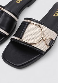 Chanclas negras con una correa trenzada beige, que cuentan con una hebilla y acentos metálicos dorados, suela lisa y diseño de punta abierta.