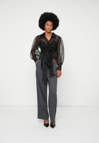 Blouse noire transparente avec motif à pois, nouée à la taille, associée à un pantalon large gris et des escarpins noirs à talons pointus.