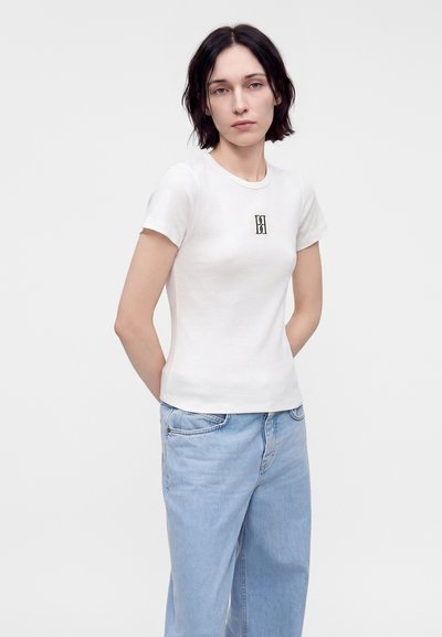 Femme aux cheveux courts et foncés portant un t-shirt blanc ajusté avec un petit logo noir et un jean bleu clair ample, debout, les mains dans le dos.