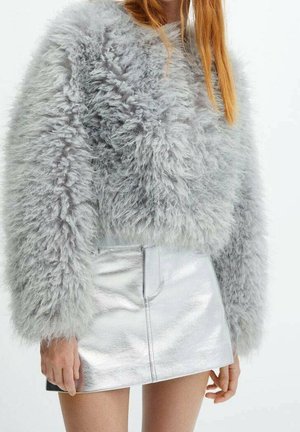 Mujer que lleva un suéter gris peludo de manga larga y una minifalda metálica plateada brillante, con cabello rojo liso parcialmente visible.