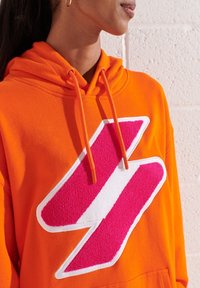 Sudadera hecha de suave tejido naranja con un gran diseño de rayo texturizado en color rosa contorneado en blanco en la parte frontal. Cuenta con capucha ajustable.