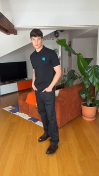 Νέος άνδρας με μαύρο πόλο και τζιν στέκεται στο σαλόνι με καφέ καναπέ, μεγάλο πράσινο φυτό, τηλεόραση και ξύλινο δάπεδο.