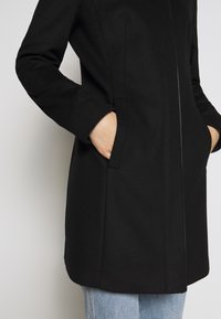 Black coat featuring a tailored design with a smooth texture, side pockets, and a straight cut. Paired with light blue jeans.