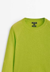 Pull tricoté vert vif avec col rond côtelé et manches longues, montrant l’étiquette du vêtement avec les détails de la marque et du matériau.