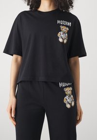 Kvinna iförd svart Moschino-outfit med teddy-björn grafik och vit text på bröstet och övre låret, stående mot vit bakgrund.