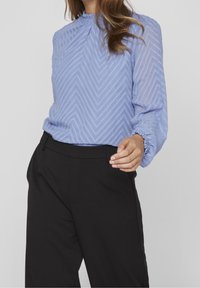 Blouse bleu clair avec un motif texturé en zigzag, un col rond et des poignets élastiques, associée à un pantalon noir ajusté.