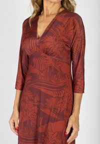 Kobieta nosząca sukienkę w kolorze rdzy z naturalnym wzorem, dekoltem w kształcie litery V i rękawami ¾, ukazująca ręce z czerwonym maniurem.
