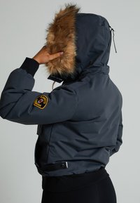 Pelēks izolēts mētelis ar kažokādas odere kapuci, ribotām aprocēm un uzšuvumu uz piedurknes. Satur pieguļošu siluetu un sānu kabatas.