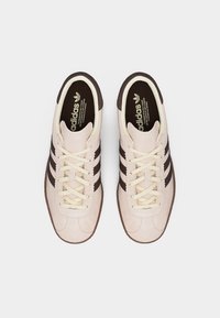 Een paar beige Adidas sneakers met bruine strepen en crèmekleurige veters, gezien van bovenaf op een witte achtergrond.