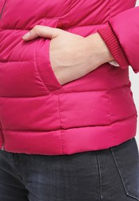 Dłoń spoczywa w kieszeni jaskrawo różowej pikowanej kurtki noszonej z ciemnymi dżinsami.