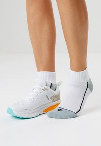 Valkoinen urheilushoe, jossa on turkoosinvärisiä ja oransseja yksityiskohtia, hengittävästä verkkomateriaalista valmistettu päällinen, yhdistettynä valkoiseen ja harmaaseen nilkkasukkaan, jossa on musta viiva.