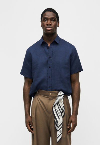 Άνδρας που φοράει μπλε σκούρο κοντομάνικο πουκάμισο μπουρνιασμένο σε παντελόνι ψηλόμεσο καφέ, με μια ασπρόμαυρη ριγέ εσάρπα κρεμασμένη από τη μέση.