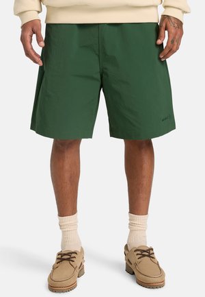 Mand iført mørkegrønne knælange shorts, cremefarvede sokker, beige snørebåndssko og cremefarvet langærmet top med synlige tatoverede hænder.