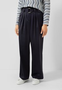 Wijd uitlopende, marineblauwe broek met een gladde textuur, voorzien van plooien en een zilveren D-ring riem. Gecombineerd met witte sneakers en een gestreept shirt.