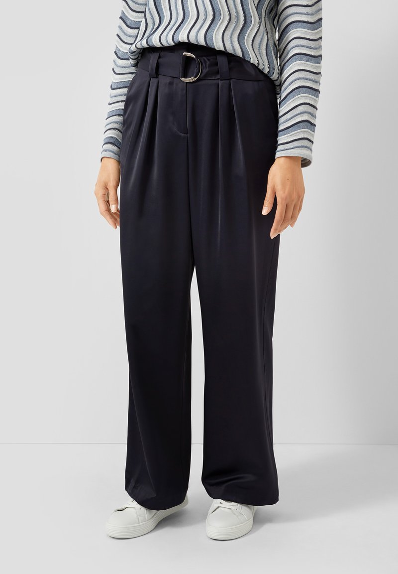 Wijd uitlopende, marineblauwe broek met een gladde textuur, voorzien van plooien en een zilveren D-ring riem. Gecombineerd met witte sneakers en een gestreept shirt.