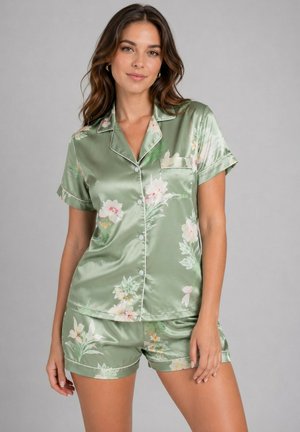 Femme portant un haut de pyjama en satin vert clair à motif floral à manches courtes et boutons, avec un short assorti, debout devant un fond gris uni.