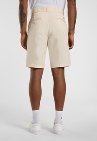 Beige Stoffshorts mit maßgeschneidertem Schnitt, ausgestattet mit zwei Gesäßtaschen und einer glatten Textur. Getragen mit weißen Schuhen und Socken.