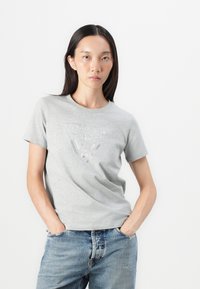 Grå bomulls t-skjorte med et trekantet rhinestones-design med "GUESS" tekst. Kortermet og med rund hals, kombinert med lyseblå denim jeans.