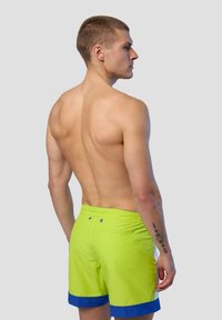 Costumi da bagno verde neon con orlo blu, caratterizzati da una texture liscia e una tasca sul retro. La vista posteriore mostra una silhouette slim e dettagli minimi.