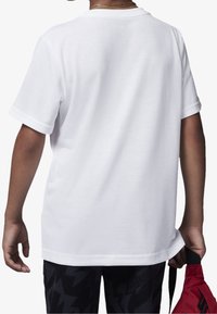 Vit bomull t-shirt med kort ärm och rund hals, med en mjuk yta. Svart mönstrad byxa delvis synlig. Röd väska hålls i handen.