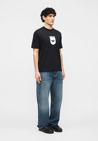 Άνδρας με σγουρά μαλλιά φοράει μαύρο μπλουζάκι με στάμπα, φαρδιά μπλε τζιν και μαύρα αθλητικά παπούτσια, στέκεται μπροστά σε απλό λευκό φόντο.