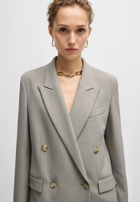 Dobbeltbrystet grå blazer lavet af tekstureret stof med en hakkrave, der har overdimensionerede knapper og en enkelt lomme på venstre side.
