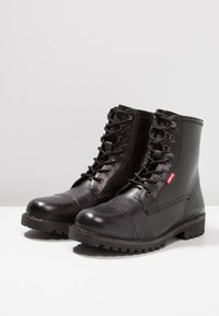 Bottes basses en cuir noir avec bouts ronds, lacets et semelle en caoutchouc texturée. Présentent une étiquette logo Levi's rouge sur le côté. Finition lisse.