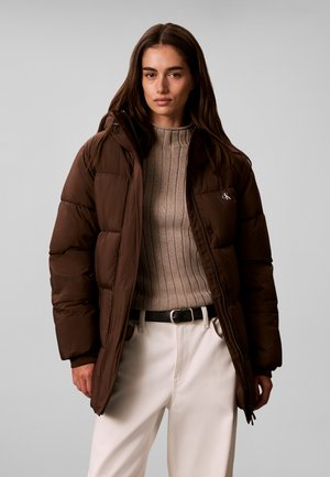 Kvinde med langt hår iført en brun dunjakke, beige ribstrikket sweater, hvide bukser og sort bælte, stående foran en ensfarvet grå baggrund.