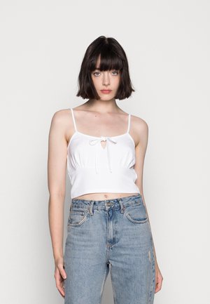 Top cropped branco com alças ajustáveis, detalhe em formato de buraco e um laço na frente. Combinado com jeans azul de cintura alta.