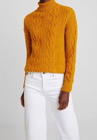 Senfgelbes Strickpullover mit hohem Rollkragen, mit einem Zopfmuster und gerippten Bündchen, kombiniert mit weißen weit geschnittenen Jeans.