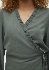 Haut portefeuille vert en tissu texturé avec un bord froncé le long de l'encolure et un détail noué sur le côté. Manches longues avec une coupe décontractée.