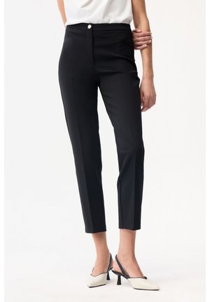 Nainen, joka pukeutuu mustiin, kapealanteisiin nilkkahousuihin ja valkoisiin slingback-korkokenkiin, kuvattu vyötärön alapuolelta, seisoo toinen käsi taivutettuna.