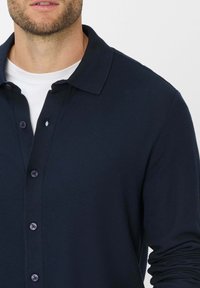 Marineblauw overhemd met kraag van textuurstof, knoopsluiting aan de voorkant en lange mouwen; heeft een witte binnenkraag.