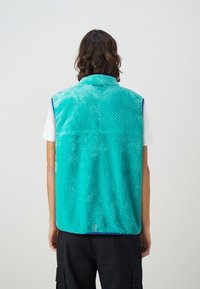Turkis fleecevest med et teksturert mønster, krage-design og sidelufter. Båret over en hvit skjorte, vist fra baksiden.