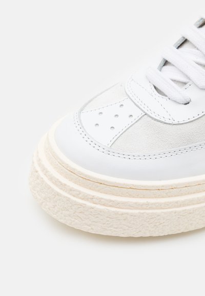 Hvit lær- og semsket skinn-sneaker med perforerte detaljer, flate lisser og en teksturert off-white plattformsåle. Minimalistisk design.