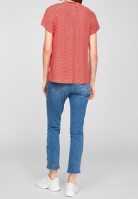 Haut rouge à manches courtes avec un motif géométrique, détail de plis au dos, associé à un jean bleu clair et des baskets blanches.