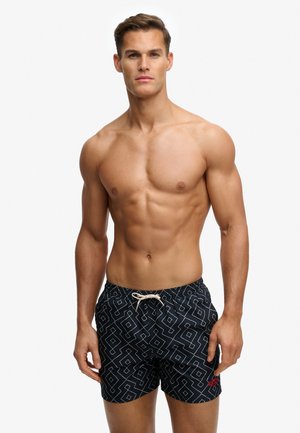 Hombre musculoso con pantalones cortos de baño negros con patrones geométricos blancos y un cordón blanco, de pie frente a un fondo liso.