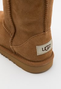 Brązowy zamszowy botek za kostkę z przeszywanym szwem, gumową podeszwą i małą beżową metką z logo "UGG". Gładka faktura z klasycznym wzorem.