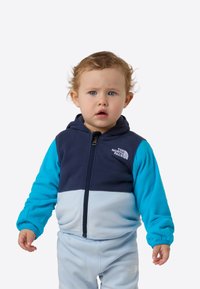 Geacă cu fermoar din fleece pentru copii mici, cu panouri albastru închis și albastru deschis, glugă și logo-ul The North Face pe piept. Textură moale.