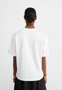 Hvid bomulds t-shirt med en afslappet pasform, korte ærmer, en rund hals og en lige kant. Har diskret branding på venstre ærme.