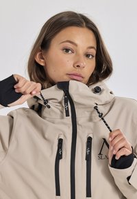 Beige wasserdichte Jacke mit schwarzen Akzenten, verstellbarer Kapuze und mit Reißverschlüssen versehenen Taschen. Glatte Textur mit Logodetails auf der Brust und an den Reißverschlüssen.