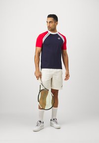 Maglietta da tennis con maniche corte rosse, corpo blu navy, dettagli bianchi e un logo. Abbinata a pantaloni corti bianchi e con una racchetta da tennis in mano.