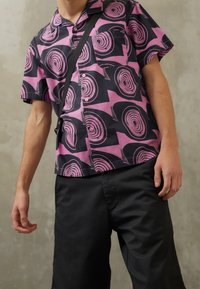 Lyhythihainen nappipaita vaaleanpunaisesta ja mustasta kankaasta, jossa on pyörteilevä kuvio. Paita on yhdistetty tummiin shortseihin ja olkalaukkuun.